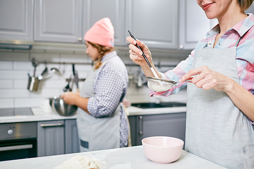 Smiling Caucasian woman sifting flour through sieve while her female colleague whisking eggs in blurred background