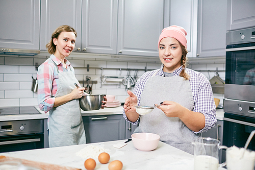 Portrait of young Caucasian female pastry cooks smiling at camera while sifting flour and whisking eggs for cake