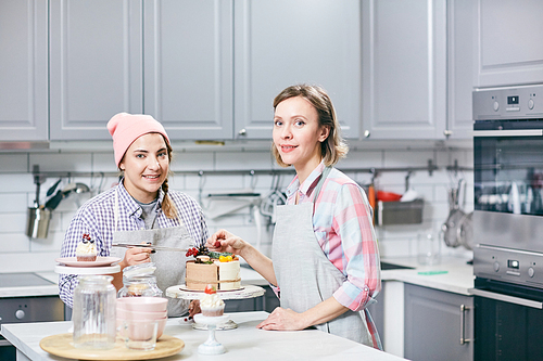 Portrait of attractive Caucasian women standing in kitchen near beautifully decorated appetizing cake and smiling at camera