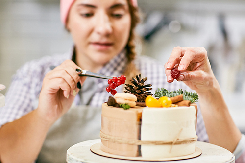 Defocused young female pastry cook decorating tasty cake with fir twigs, cones, currants and raspberries, close-up view