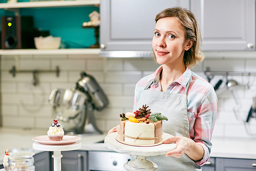 Portrait of attractive Caucasian woman standing in kitchen near decorated appetizing cake and smiling at camera