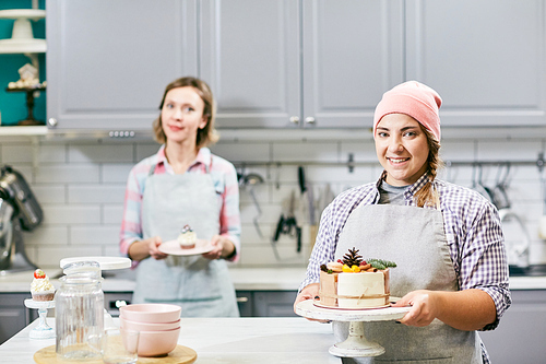 Portrait of young pretty women in aprons standing in kitchen with delicious cakes and smiling at camera happily