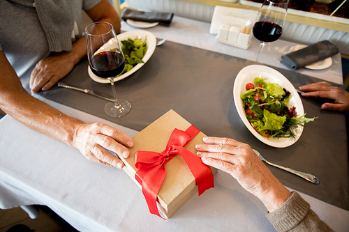 Top view of unrecognizable senior couple tenderly joining hands while exchanging presents at table in cafe, copy space