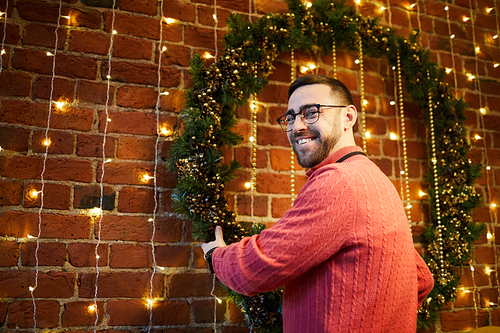 Happy man in pink pullover hanging big decorative coniferous wreath on office wall with sparkling fairy lights