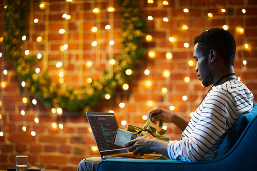 African-american it-engineer untying golden ribbon on gift-box on Christmas eve while sitting in office