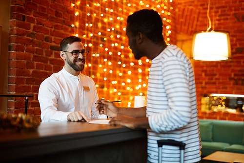 Happy hotel receptionist talking to foreign traveler by counter and helping him to register