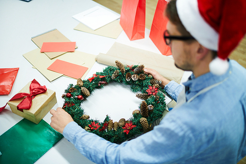 Busy young man standing at table with gift box and colorful papers and decorating Christmas wreath in modern workshop