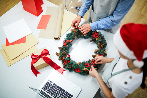 High angle view of unrecognizable busy designers standing at table with craft paper, gift box and laptop and making Christmas wreath together in holiday home decor workshop