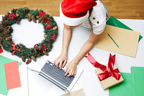 High angle view of busy woman in Santa hat leaning on big table with Christmas decorations and typing on laptop in workshop
