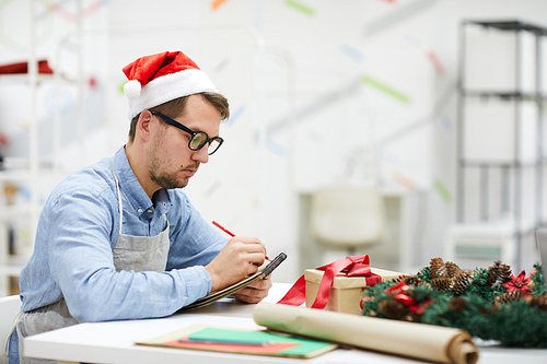 Serious pensive male designer in Santa hat and glasses sitting at table and making notes in sketchpad while working in crafting studio, he planning work and composing list of orders