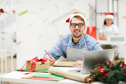 Happy handsome creative designer with beard wearing Santa hat and glasses sitting at messy table and smiling at camera in office, he making sketch