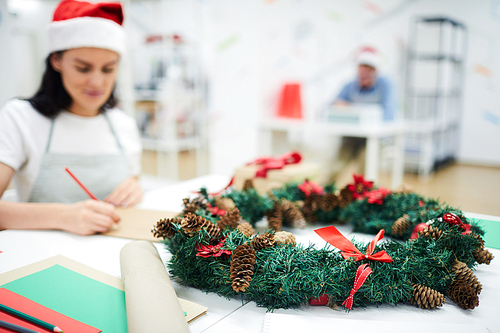 Close-up of beautiful Christmas wreath with ribbons and pine cones place on office table, woman drawing in background