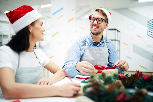Happy positive young colleagues in Santa hats and aprons sitting at table and talking while packing Christmas gifts in office