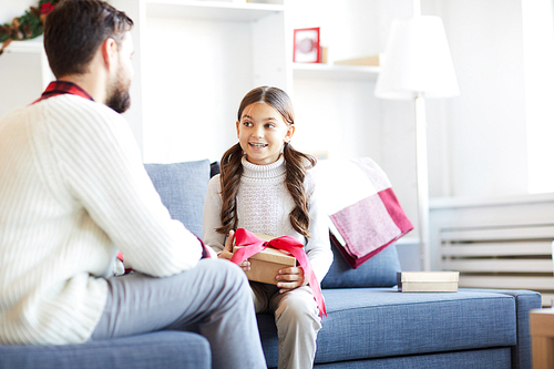 Little girl with giftbox sitting on sofa with her dad and talking to him in living-room
