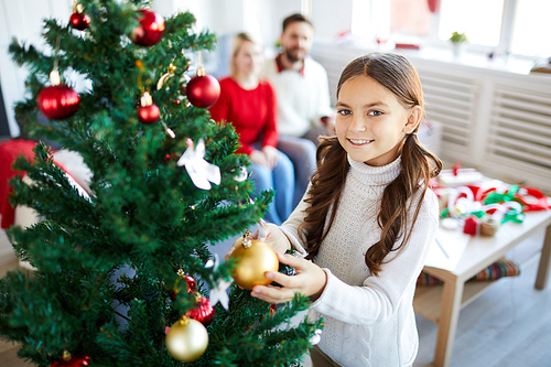 Pretty little girl decorating xmas tree with golden and red toy balls while looking at you