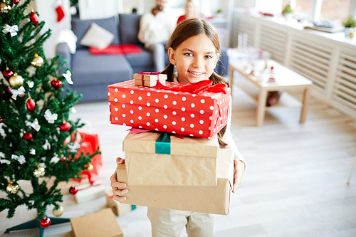 Little girl showing you stack of Christmas gifts while standing by decorated firtree