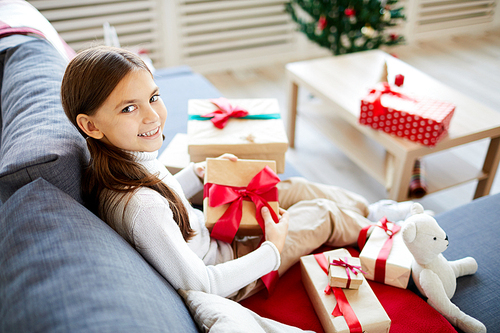 Little girl looking at you while sitting on sofa and looking through her xmas gifts