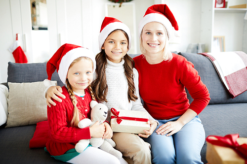 Two cute little girls and their mother in Santa caps sitting on sofa at home