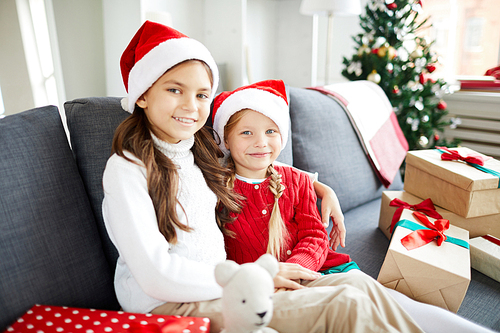 Cute happy little girls in Santa caps and casualwear sitting on sofa in living-room