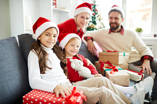 Little girls and their parents in Santa caps sitting on sofa with heap of gifts on xmas eve