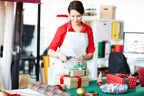 Young woman in apron standing by table and cutting ribbon on top of giftbox with xmas present