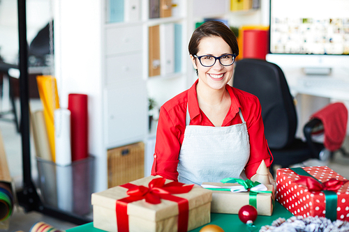 Cheerful young female in red blouse and apron looking at you while sitting by table with gifts