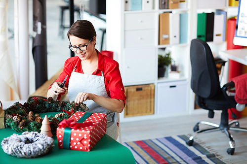 Creative young woman in workwear standing by table with xmas staff and finishing up with decorative wreath
