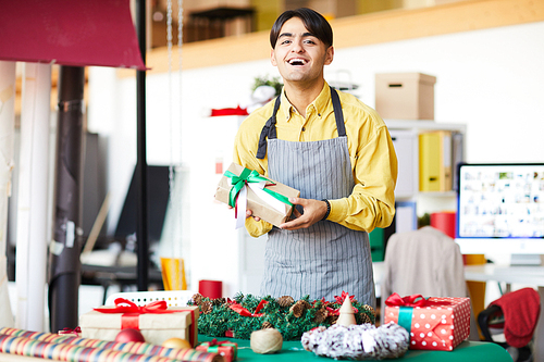 Young laughing man in apron standing by table with heap of xmas stuff in his studio