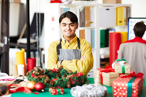 Happy young cross-armed man looking at you while standing by table with xmas gifts and decorations