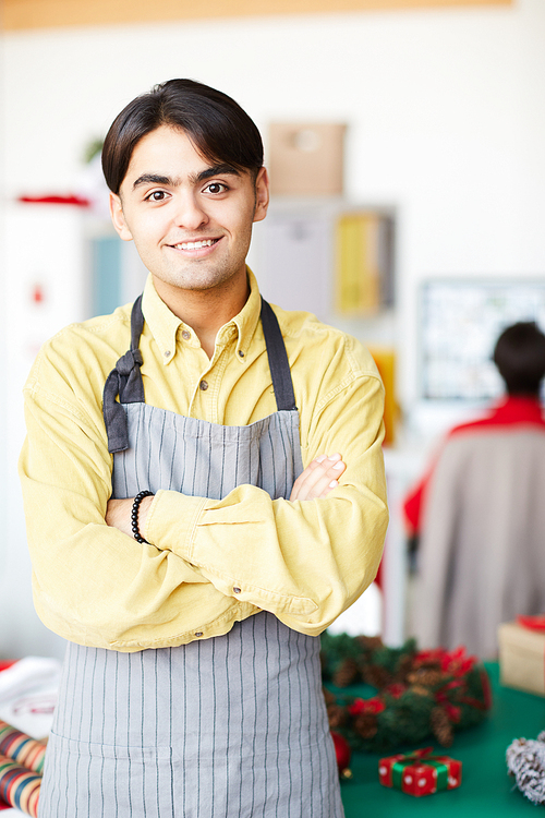 Young smiling man in apron crossing his arms on chest in studio of design with colleague on background