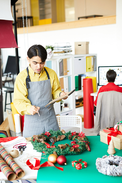 Young creative man with notepad and pencil standing by table with xmas stuff and pointing at wreath