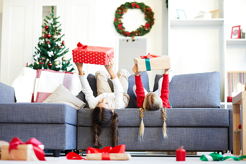 Two little girls lying on sofa and holding giftboxes while stretching arms in front of themselves