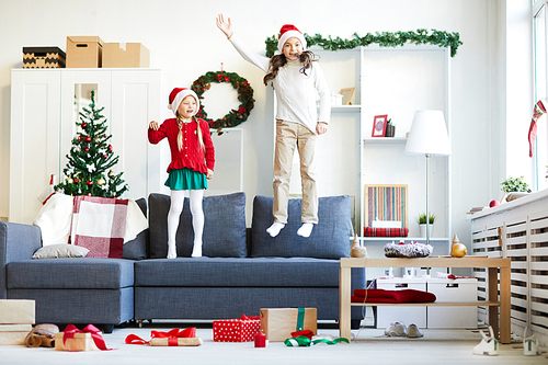Two playful girls in Santa caps and casualwear leaping on sofa on Christmas evening