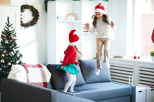 Two cute Santa girls jumping on sofa while having fun at home on xmas evening