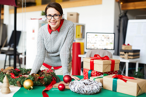 Happy woman in grey knitted sweater leaning over table with Christmas decorations in studio