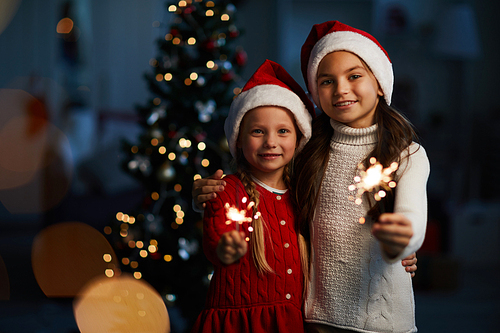 Two little Santa girls with burning bengal lights looking at you on background of decorated firtree