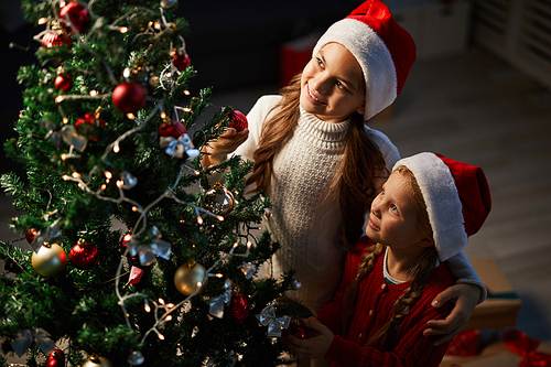 Two little sisters in Santa caps looking at decorated xmas tree on holy night