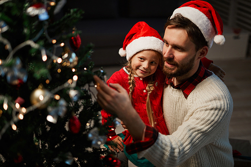 Young man and his little daughter decorating Christmas tree with toys and garlands