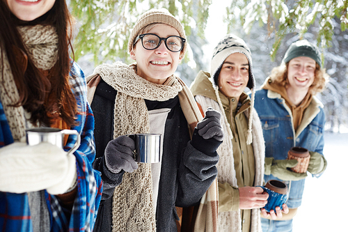 Group of young people smiling cheerfully at camera while posing in winter forest standing in row, focus on young woman holding metal cup