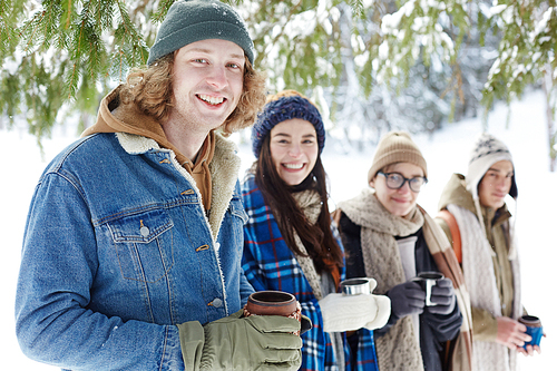 Group  friends smiling cheerfully at camera while posing in winter forest standing in row, focus on young man holding cup in front