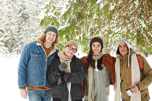 Waist up portrait of group of happy young people posing in winter resort standing under fir tree in beautiful snowy forest