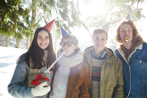 Group of four young people celebrating Christmas outdoors in beautiful forest, all wearing party caps