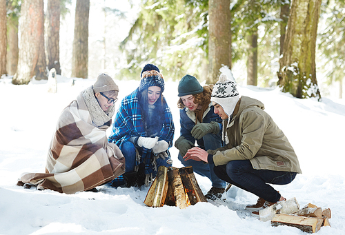 Full length portrait of four young people camping in winter forest sitting in circle round fire