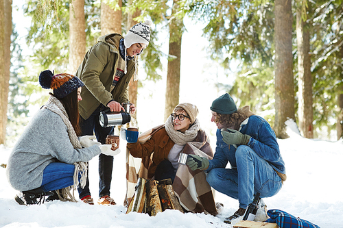 Full length portrait of four young  friends camping in winter forest sitting in circle round fire and pouring hot drinks