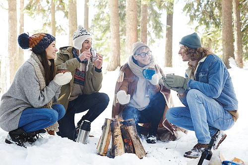 Full length portrait of four young people camping in winter forest sitting in circle round fire and chatting while drinking hot cocoa