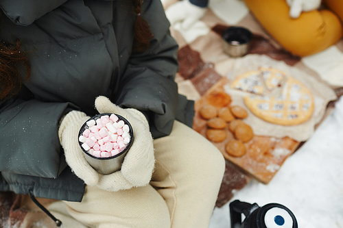 Close-up of unrecognizable woman in warm mittens sitting on ground and drinking delicious cocoa with marshmallows outdoors