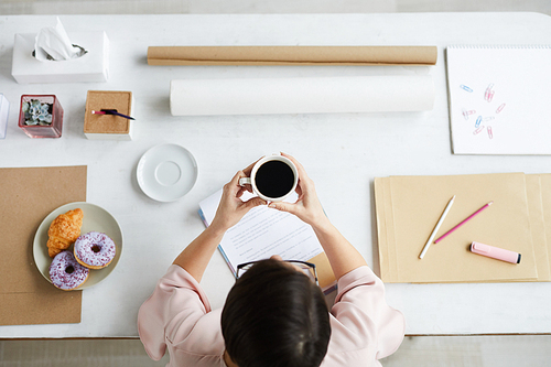 Contemporary office worker with cup of espresso sitting by desk with office supplies around