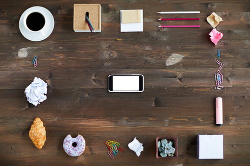 Creative arrangement of modern smartphone surrounded by stationery, crumpled paper, pastry and cup of coffee laid out on wooden desk