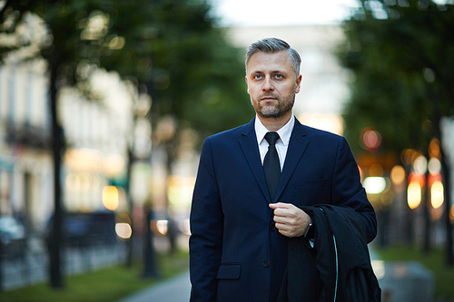Mature serious businessman in elegant suit holding his coat while walking in urban environment