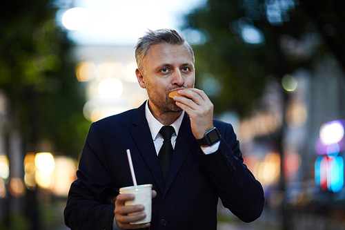 Contemporary businessman or director in suit eating quickly his sandwich outdoors while hurrying for briefing